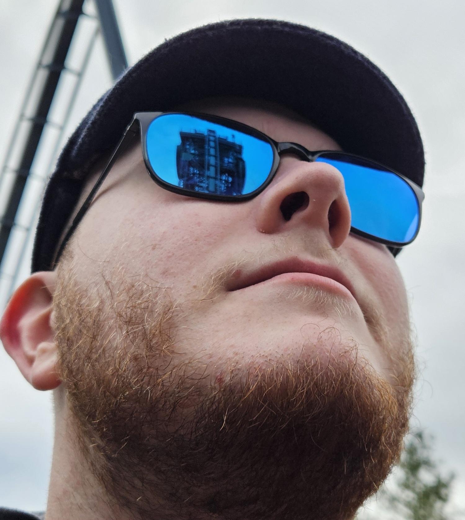 Sid, a man with sunglasses, a peaky blinders cap and a Rohirrim lord of the rings sweater is staring at the rollercoaster: Baron 1898 in themepark the Efteling. In the reflection of the sunglasses he is wearing you can see the iconic drop that is at a 87 degrees angle.